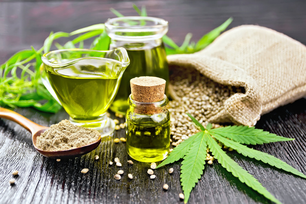 Hemp oil in two glass jars and sauceboat with grain in the bag, leaves and stalks of cannabis, a spoon with flour on the background of wooden boards